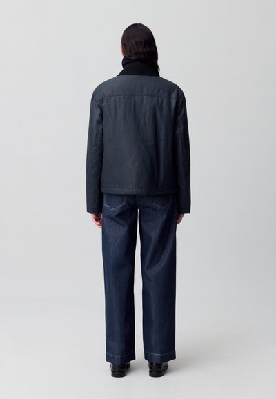 Veste noire avec un col haut et un tissu texturé, associée à un pantalon en denim bleu à jambes larges avec des coutures contrastées et une coupe droite.