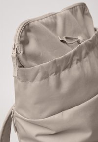 Beige stofrygsæk med en lynlåslomme øverst og en snørelukning inde i hovedrummet.