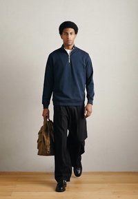 Tmavomodrá mikina na zips, čierne široké nohavice, lesklé čierne topánky, držiaci hnedú kabelku, stojaci na drevenej podlahe s jednoliatym pozadím.