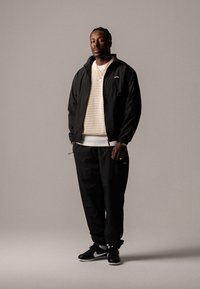Jachetă neagră din nailon peste un pulover ușor, tricotat, purtată cu pantaloni negri și adidași negri Nike. Design simplu, fără modele vizibile.