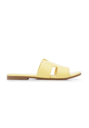 Sandalo piatto giallo con design traforato e suola marrone, mostrato di lato su sfondo bianco.