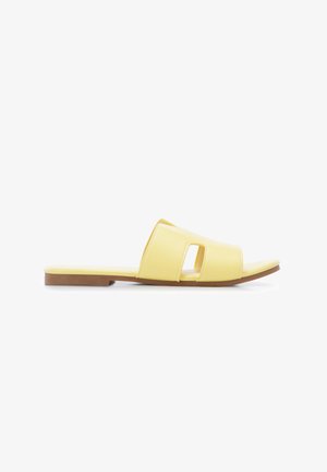 Gele platte slide-sandaal met uitgesneden ontwerp en bruine zool, van opzij getoond op een witte achtergrond.