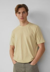 Lichtgele t-shirt met korte mouwen in een zachte textuur en een ronde halslijn, gecombineerd met olijfgroene broek, handen in de zakken tegen een neutrale achtergrond.