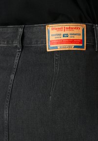 Svart denimbukse med en rektangulær lys brun etikett som leser Diesel Industry, registrert siden 1978, garantert, jeans av overlegen kvalitet.