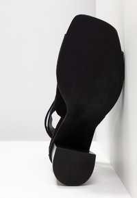 Sandales à talons noirs avec une tige en daim, bout rond et talon épais. La semelle est lisse et texturée avec des indications de taille.