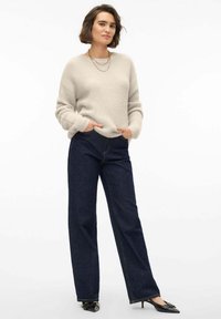 Maglione beige fluffy e jeans a gamba larga blu scuro. Il maglione ha un collo a giro e maniche lunghe. I jeans hanno cuciture visibili e tasche anteriori.