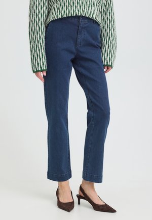 Vrouw met rechte blauwe spijkerbroek, groen-wit gebreide trui met patroon en donkerbruine puntige slingback hakken, staand op een witte vloer.
