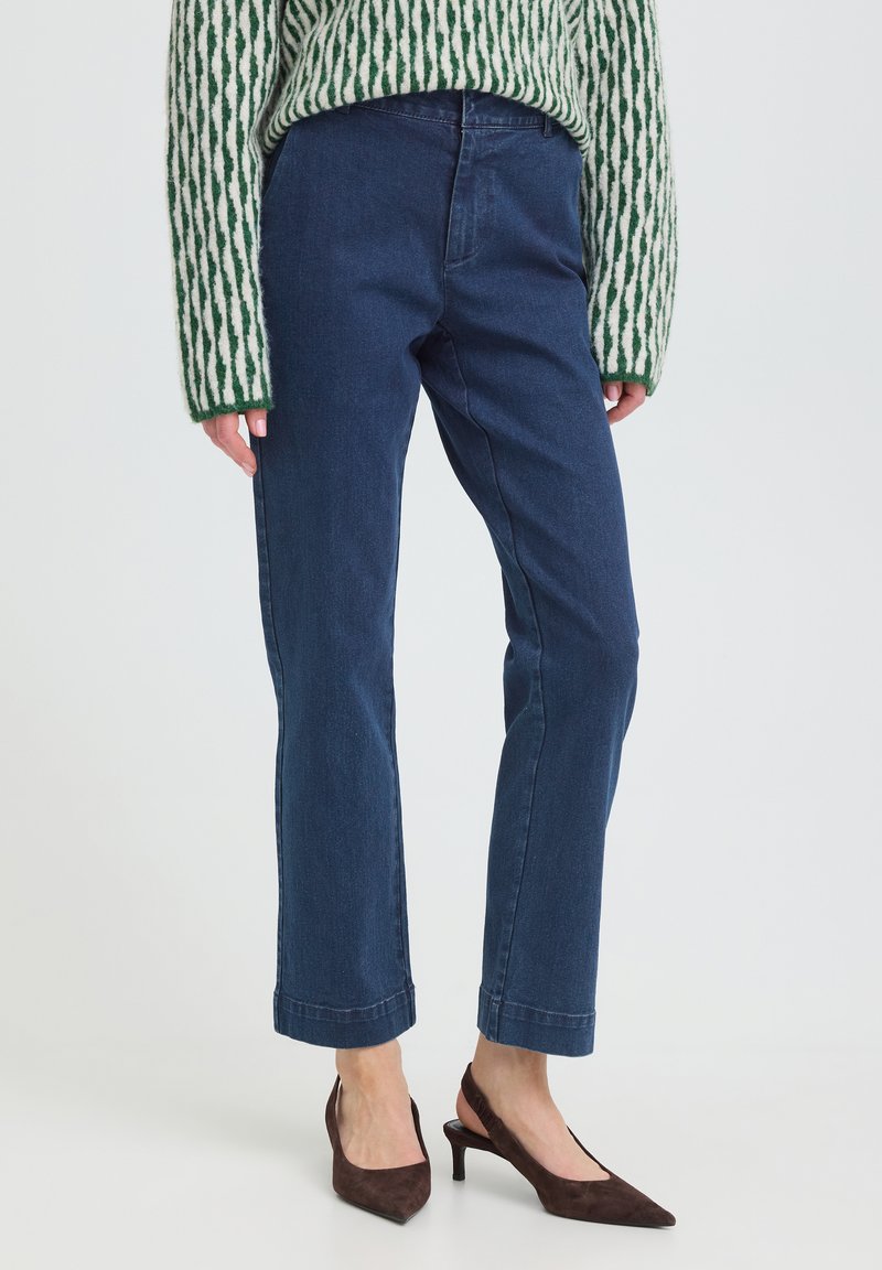 Femme portant un jean droit bleu, un pull vert et blanc à motifs, et des talons slingback pointus marron foncé, debout sur un sol blanc.