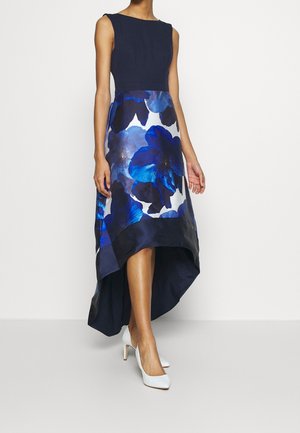 Vrouw draagt een mouwloze navy jurk met blauwe bloemenrok en hoog-laag zoom, gecombineerd met witte hoge hakken, staand tegen een effen muur.