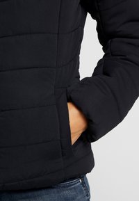 Main dans la poche latérale d'une veste noire matelassée portée avec un jean en denim bleu, sur un fond gris clair uni.