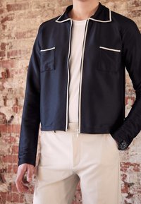 Man i svart zip-up jacka med vit kant, vit skjorta, beige byxor och svart armbandsur står mot en texturerad tegelfond.