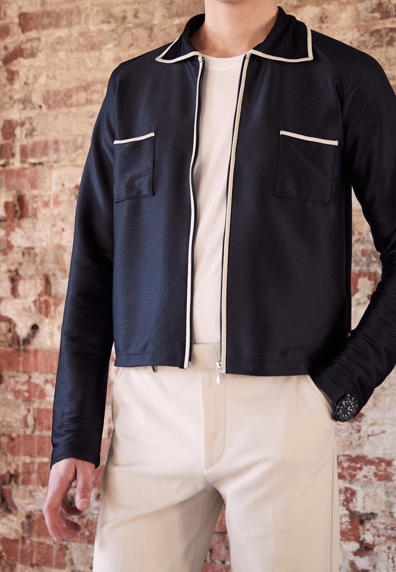 Man i svart zip-up jacka med vit kant, vit skjorta, beige byxor och svart armbandsur står mot en texturerad tegelfond.