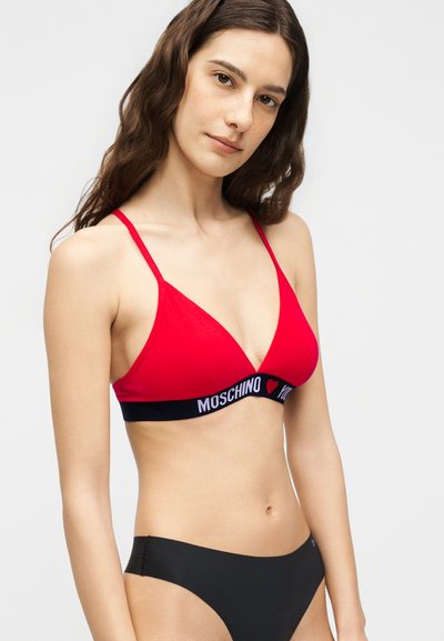 Γυναίκα που φοράει κόκκινο τριγωνικό σουτιέν με την επιγραφή "Moschino" και μαύρο κάτω μέρος μπικίνι, ποζάρει μπροστά σε απλό φωτεινό φόντο.
