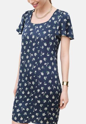 Femme portant une robe à manches courtes bleu marine avec un imprimé floral de marguerites blanches, un collier chaîne en or et un bracelet manchette en or.