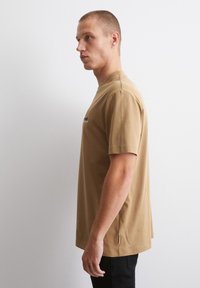 Brun bomull t-skjorte med rund hals, korte ermer og sidespalte detaljer, med minimal svart tekst på brystet.