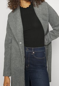 Donna che indossa un cappotto grigio a spina di pesce sopra un top nero a coste e jeans scuri a vita alta, con una mano sulla vita, su uno sfondo neutro.