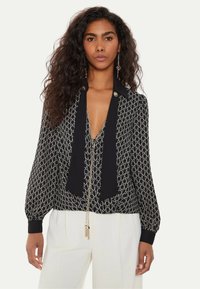 Donna con capelli lunghi e ricci, che indossa una blusa nera a motivo con scollatura profonda e pantaloni bianchi a vita alta, rivolta verso l'avanti.