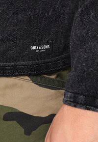 Czarny sweter dzianinowy o tkanym wykończeniu, z metką informującą "ONLY & SONS EST. MMXIII", zestawiony z spodniami w kamuflażowy wzór.