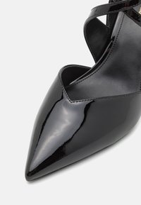 Chaussure noire brillante à bout pointu avec un design à brides croisées et une finition en cuir lisse sur un fond clair.