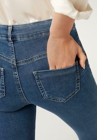 Mano metida en el bolsillo trasero de unos vaqueros de mezclilla azules, usados con una camisa blanca sobre un fondo claro.