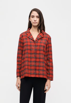Modèle portant une chemise rouge à carreaux boutonnée et un pantalon noir, debout devant un fond uni clair.