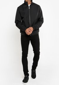 Sort bomberjakke med høj krave, sølv lynlås og ribbede manchetter. Sammen med tætsiddende sorte jeans og sorte ruskinds sneakers.