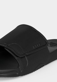 Sandale slide noire réglable avec semelle texturée et logo "REEF" embossé sur la large bride.