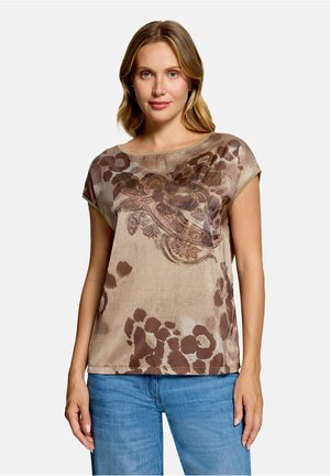 Femme aux cheveux blonds portant un haut beige à manches courtes avec des motifs floraux marron et paisley et un jean bleu, debout devant un fond uni.