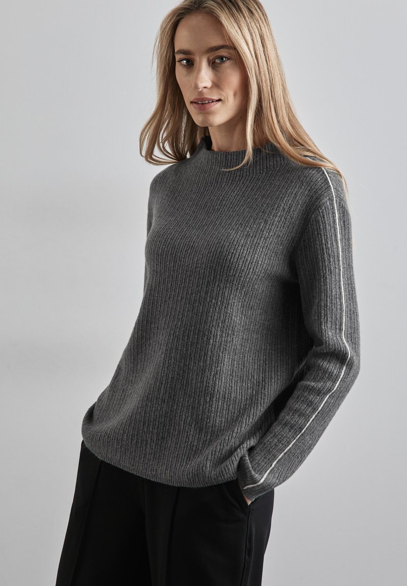 Grauer gerippter Pullover mit hohem Kragen und weißen Seitenstreifen. Lange Ärmel, weiche Textur, lockere Passform, kombiniert mit schwarzen Hosen.
