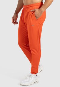 Orange atletiska byxor med elastisk midja, dragsko och sidofickor. Slät tyg med en åtsittande design och subtil märkning.