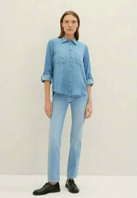 Nais is wearing a kerge sinine denim särk, mille varrukad on üles keeratud, sellele sobivad helesinised teksad ja mustad nahkkingad, seistes ühevärvilise tausta vastu.