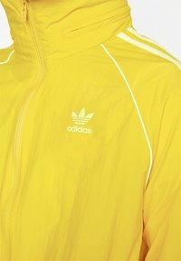 Žlutá lehká bunda na zip s bílými akcenty a logem Adidas na levé části hrudi. Texturovaná tkanina, sportovní design.