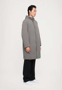 Doudoune grise avec capuche, dotée d'une fermeture à boutons et de poches latérales. Portée sur un pantalon noir et des baskets blanches, vue de profil.
