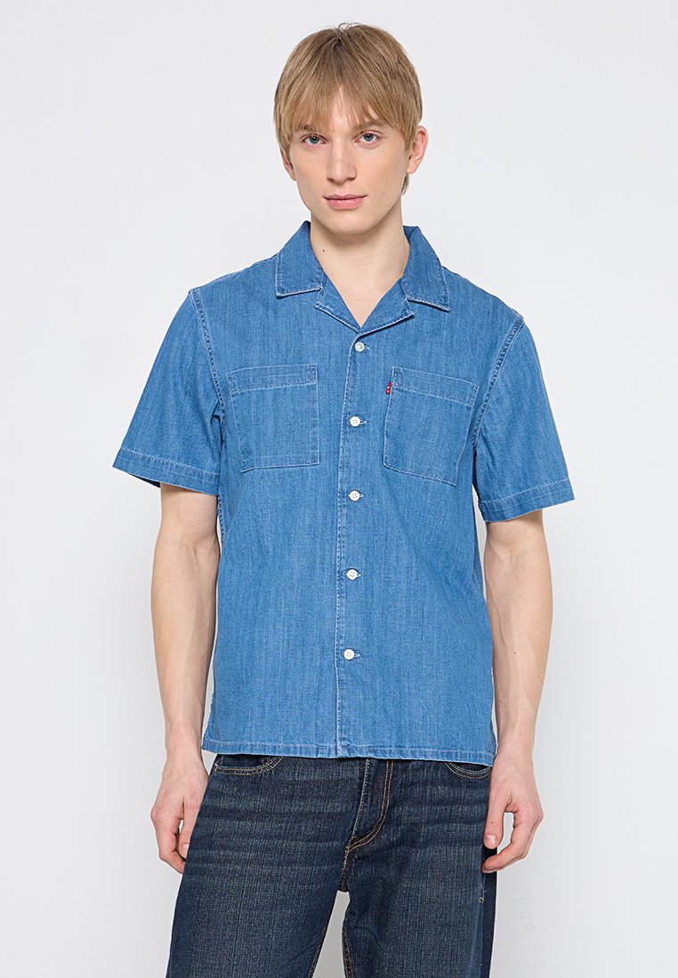 Jeune homme portant une chemise en jean bleu à manches courtes, avec deux poches poitrine, et un jean foncé, se tenant contre un fond blanc.
