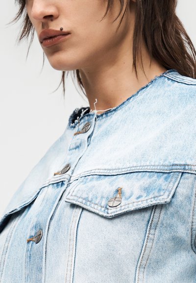 Κοντινό πλάνο ενός ατόμου που φορά ένα ανοιχτό μπλε τζιν μπουφάν με μεταλλικά κουμπιά, δείχνοντας το κάτω μέρος του προσώπου και τον λαιμό με μια μεγάλη ελιά πάνω από το χείλος.