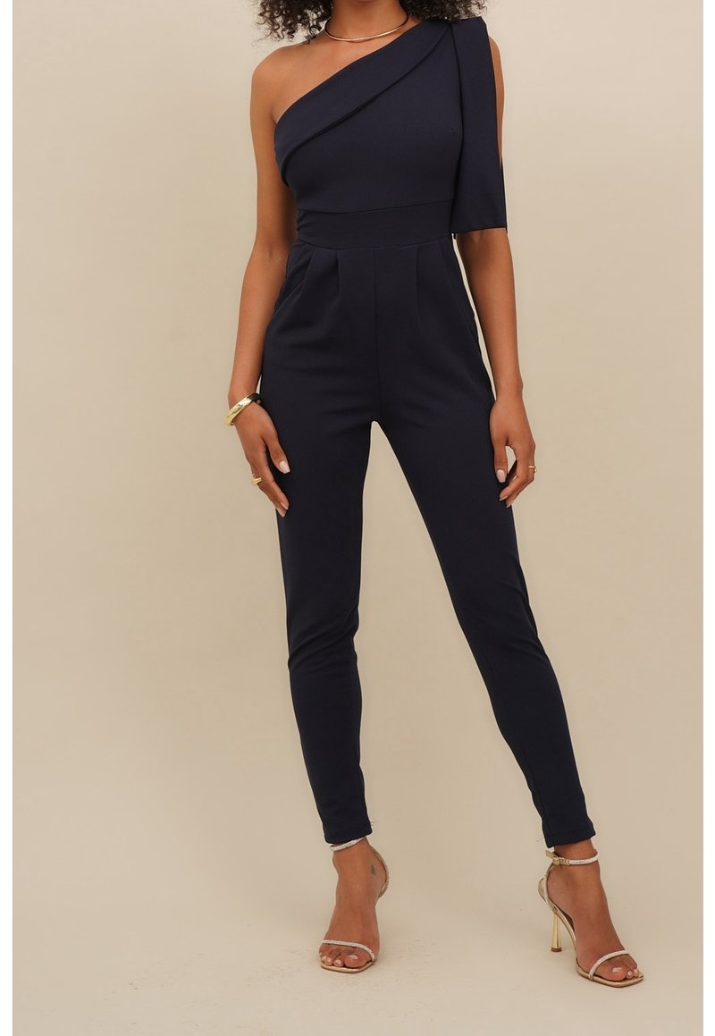 Jumpsuit blu navy a spalla unica con corpetto aderente, gambe affusolate e dettagli a pieghe; realizzato in tessuto morbido. Tacchi a spillo dorati completano l'outfit.