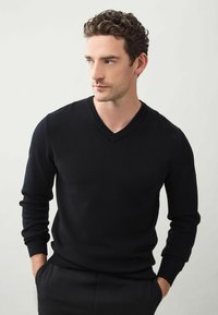 Pulover negru cu decolteu în V, dintr-un material tricotat moale, cu mâneci lungi și manșete cu nervuri, asortat cu pantaloni închiși la culoare.
