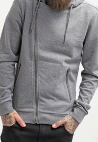 Sudadera con capucha gris con cierre de cremallera diagonal, confeccionada con tela suave, que incluye dos bolsillos laterales con cremalleras y puños acanalados.