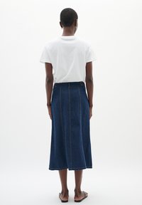 Denim midi krilo v temnem pranju, z oprijetim pasom in razširjeno silhueto. Oblačeno belo majico, z ravnimi sandalami. Preprost dizajn in šivi.