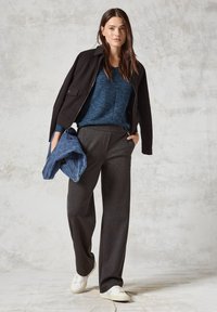 Svart jacka, blåmönstrad skjorta, grå vida byxor och vita sneakers. Håller en blå denimväska. Neutral bakgrund.