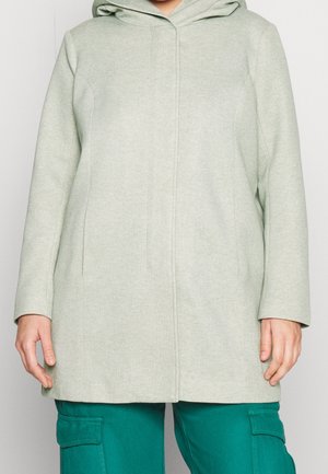 Jasnoszary płaszcz z kapturem o prostych liniach, noszony na zielonych spodniach bojówkach, pokazany od ramion do połowy ud z luźno opuszczonymi ramionami.