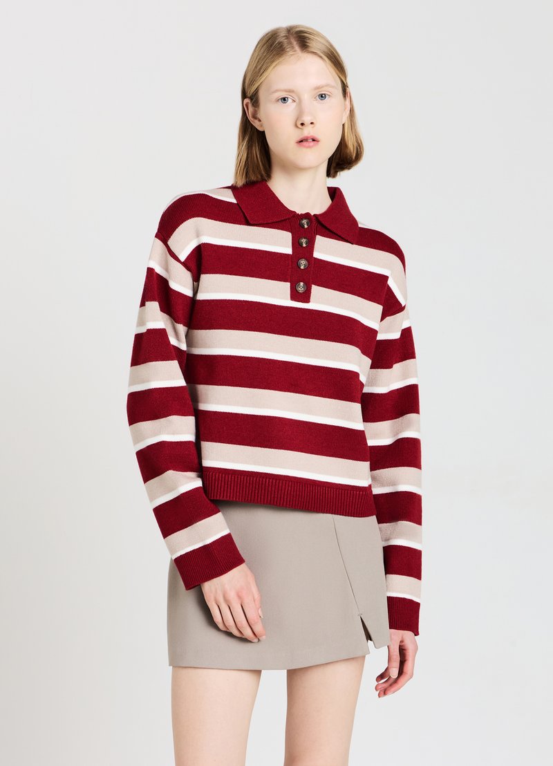 Pull rayé rouge et beige avec col polo, fermeture à l'avant à cinq boutons, poignets côtelés et longueur raccourcie. Assorti à une jupe beige.