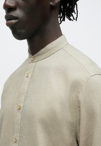 Nærbilde av en person iført en beige lin-skjorte med mandarinkrage og treknapper, som viser detaljer på nakke og skulder.