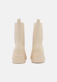 Une paire de bottes épaisses beiges à plateforme vues de l'arrière, avec des tiges hautes et des languettes de traction sur un fond blanc uni.