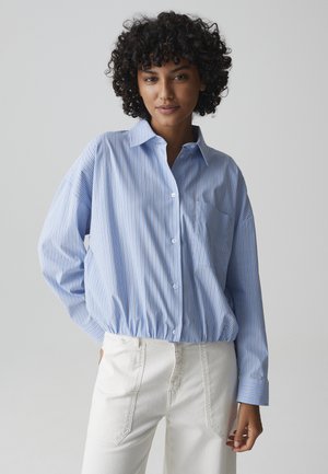 Kobieta o kręconych włosach, ubrana w niebieską koszulę w paski na guziki i białe spodnie, stoi z jedną ręką w kieszeni na szarym tle.