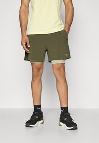 Grønne sportsbukser med lagdelt design, sidelommer og hvit aksentlogo. Svarte joggesko med grønne detaljer og mørke sokker.