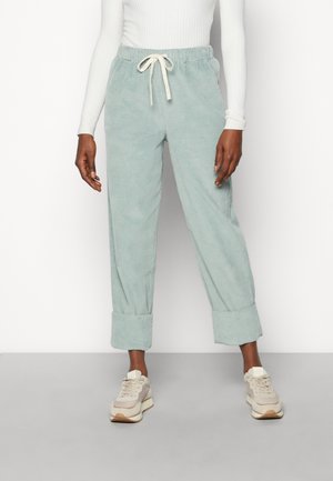 Person trägt hellgrüne Cordhose mit hochgekrempelten Bündchen, weißer Kordel, weißem Langarmshirt und beigen Sneakern und steht auf grauem Boden.