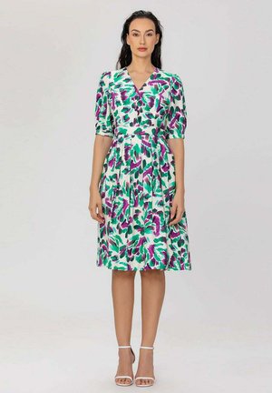 Femme debout, vêtue d'une robe blanche mi-longue à motifs floraux verts et violets, manches bouffantes et talons blancs à bride cheville.