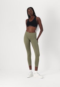 Svart sports-bh med logo og olivengrønne høyt liv leggings. Leggingsene er tettsittende, elastiske og har en glatt tekstur. Hvite sneakers.