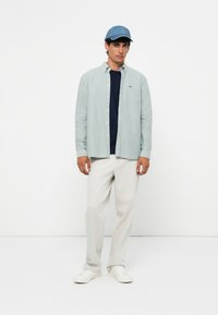 Lys grøn skjorte med knapper, marineblå T-skjorte, beige bukser og hvite joggesko. Inkluderer også en blå caps og en liten logo på skjorten.